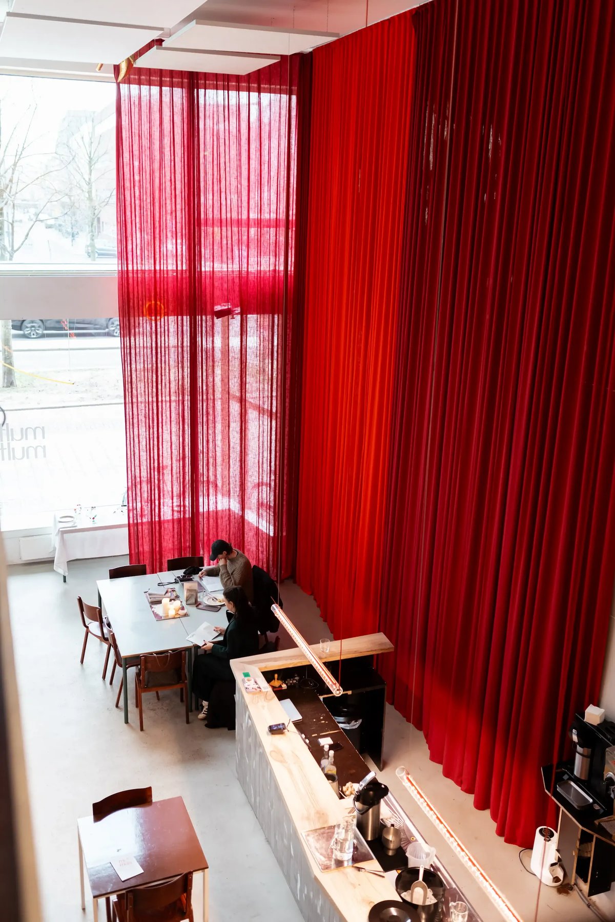 Lofty café space with dramatic red curtains, natural light, and guests working at a shared table near the window.