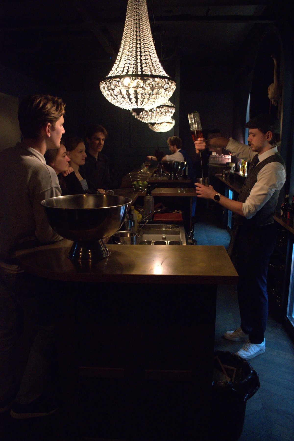 Bartender pouring spirits behind the bar at Le Flamant Bleu, with guests seated under chandeliers